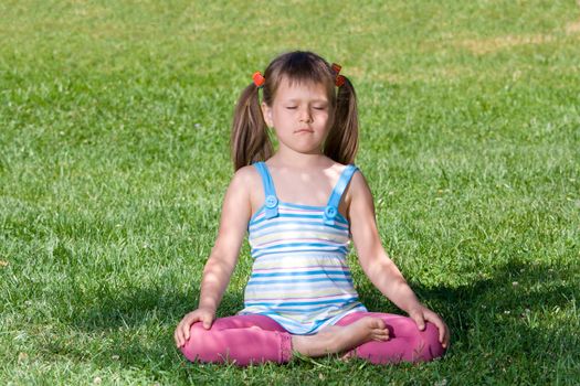 Healthy lifestyle. Little cute girl who is sitting and meditating in asana under tree shadow with closed eyes on the green grass