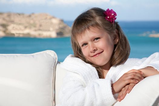 Portrait of happy smiling little girl with flowered hair in white bathrobe relaxing on terrace divan and looking at camera on the sea background