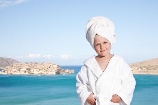 Portrait of happy smiling little girl with towel on the head in white bathrobe relaxing on terrace and looking at camera on the sea background