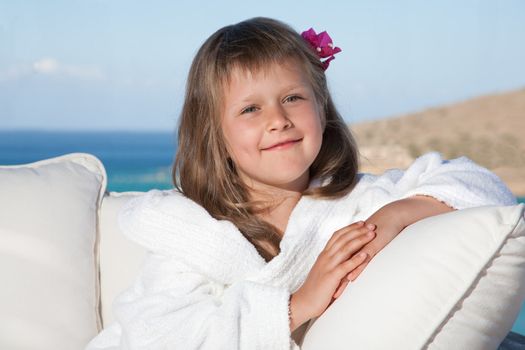 Portrait of happy smiling little girl with flowered hair in white bathrobe relaxing on terrace divan and looking at camera on the sea background