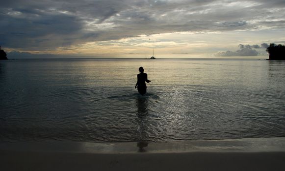 Silhouette of a woman walking into the sea at sunset with a catamaran on the horizon