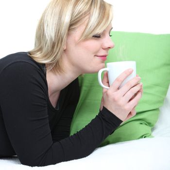 Beautiful young woman drinking coffee lying on her stomach on a couch with her eyes closed as she savours the aroma