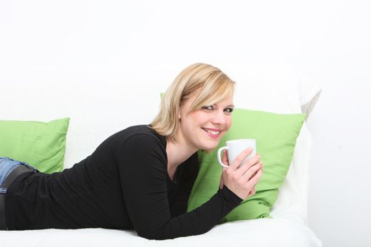 Young cheerful woman enjoying a cup of coffee as she lies on her stomach on a couch relaxing