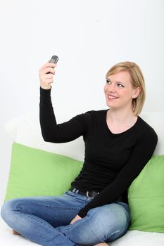Young woman smiling and posing cross-legged on a couch while photographing herself using the camera on her mobile phone