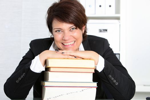 Dedicated hardworking professional woman sitting at her desk in her office with her head resting on a high stack of books smiling at the camera