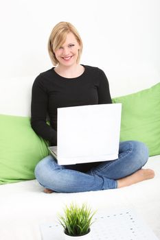 Smiling young blonde woman sitting crss legged on a couch in her living room with her laptop balanced on her lap