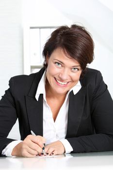 Smiling businesswoman at her desk looking up at the camera as she writes a short memo to herself