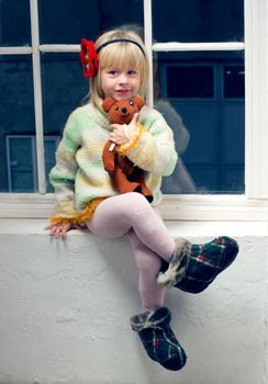 3 year old girl in knitted sweater and tights with a toy in his hand sitting on the window
