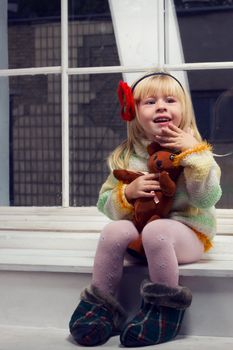 little girl in a knitted sweater and tights with a toy in his hand sitting on the window