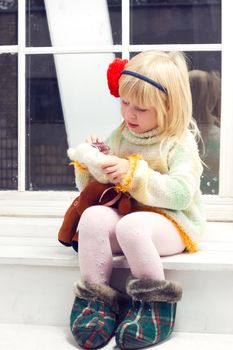 little girl in a knitted sweater with a flower and a bandage on his head with a toy in his hand sitting on the window