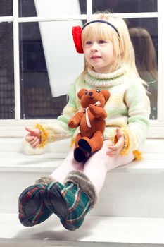 blonde little girl in knit sweater with a flower and a bandage on his head with a toy in his hand sitting on the window