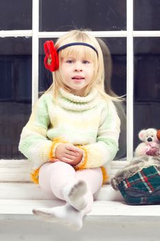 blonde little girl in knitted sweater sitting on the window and looks into the camera