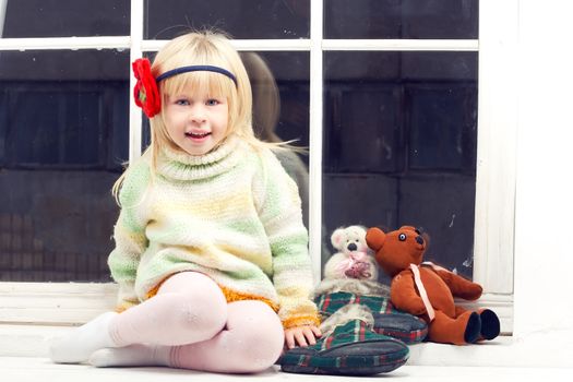 blonde little girl in knitted sweater sitting near the window of toys
