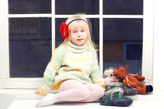 blonde little girl in knitted sweater sitting near the window of toys
