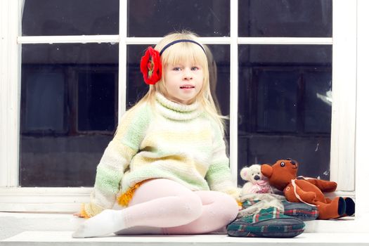 blonde little girl in knitted sweater sitting near the window of toys