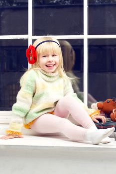 beautiful little girl blonde in a knit sweater sitting beside toys in the window and looks into the camera