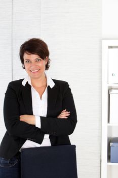 Smiling attractive female corporate executive standing behind her office chair with her arms folded