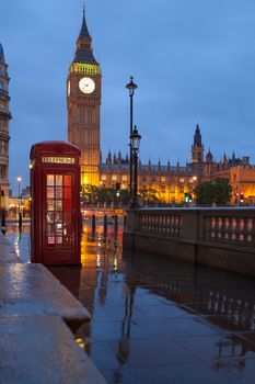Red public telephone box and illuminated clock on Big Ben tower of Westminster Palace in twilight with reflection on wet footway, Great Britain