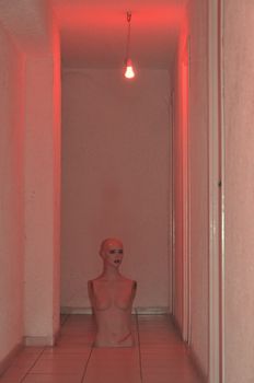 Broken mannequin doll under red light in underground corridor.