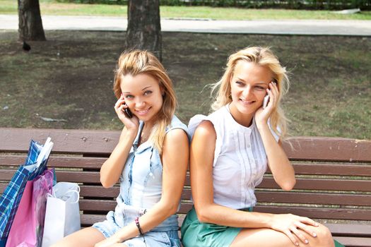 Two beautiful young women talking on the phone while sitting on a park bench