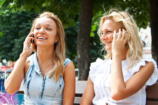 Two beautiful young women talking on the phone while sitting on a park bench