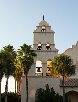 Setting sun illuminates spanish mission style church tower with bells and palm trees