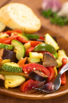 Fresh homemade Ratatouille made of eggplant, zucchini, bell pepper and tomato and seasoned with herbs (garlic, thyme, oregano) with fork and baguette on wooden plate (Selective Focus, Focus one third into the meal)