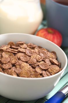 Bowl of chocolate corn flakes cereal with apple, cup of coffee/tea and a jug of milk in the back (Selective Focus, Focus in the middle of the cereal)