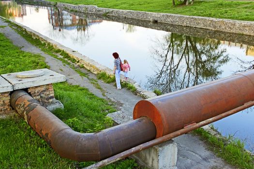 Woman and little girl going by waters bank. Large rusty metal pipe over the reservoir. View from the top