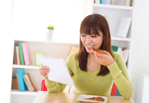 Asian woman eating pizza and looking at digital tablet computer at home