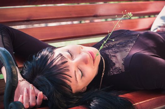 Young woman enjoying the sun, lying on a bench, with closed eyes and a flower in her lips