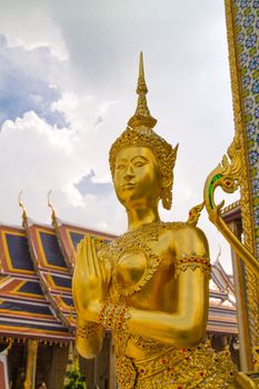 Wide angled view of Buddha sculpture Kinora or Kinnaree ( mythological creature, half bird, half man ) at Wat Phra Kaeo and Grand Palace in Bangkok, Thailand. Many details of Grand Palace in the background, also visible are beautiful cloudscape with blue sky and cumulus clouds on one sunny day in Bangkok.