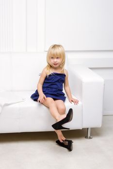 Little girl 3 years old sitting on a white sofa in her mother's dress and shoes high heels