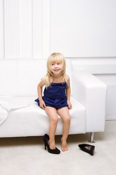 Little girl 3 years old sitting on a white sofa in her mother's dress and shoes high heels