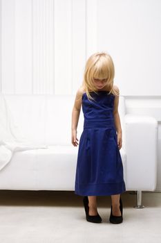 Little girl 3 years old standing near the white sofa in her mother's dress and shoes with heels