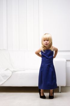 Little girl 3 years old standing near the white sofa in her mother's dress and shoes with heels