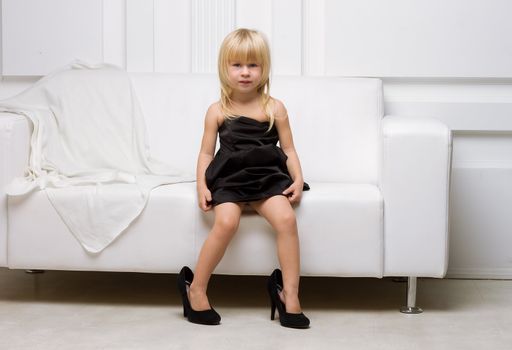 Girl 3 years old in her mother's high heels sitting on a white sofa