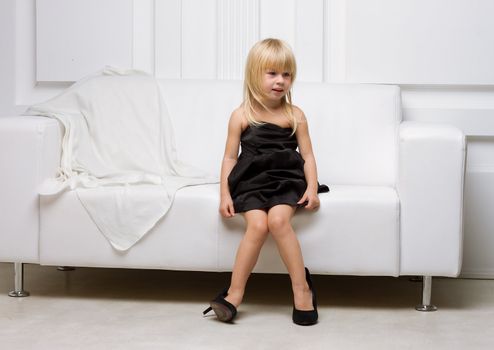 Girl 3 years old in her mother's high heels sitting on a white sofa