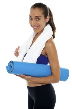 Side profile of a fit lady holding blue mat in hands and towel around her neck.