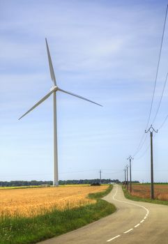 Image of a road in a field area with a row of electricity poles at the right side and a wind turbine at the left side.