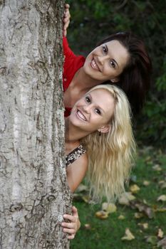 Two pretty young ladies smiling from behind a tree trunk
