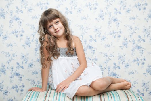Portrait of sincere cheerful tender young blond girl child with grey eyes and wavy long hair in relaxed sitting posture looking at camera