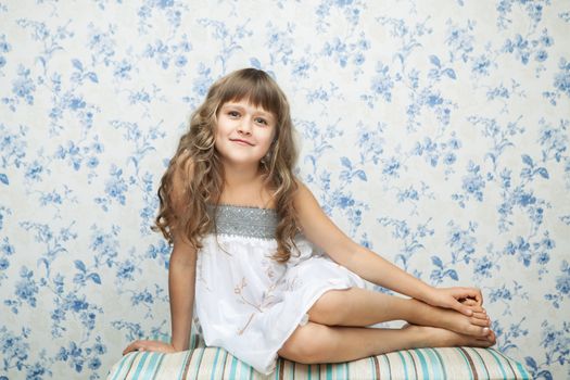 Portrait of sincere cheerful tender young blond girl child with grey eyes and wavy long hair in relaxed sitting posture looking at camera