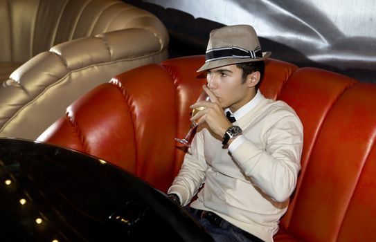 Man with hat seated on sofa in night club, with a glass of wine on the hand