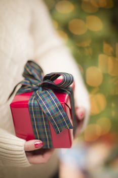 Woman Wearing A Sweater and Seasonal Red Mittens Against an Abstract Green and Golden Background Holding A Beautifully Wrapped Christmas Gift with Narrow Depth of Field.