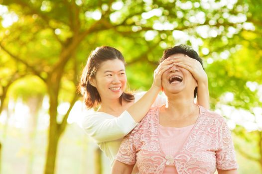 Asian adult daughter covering her senior mother eyes by hands at outdoor park