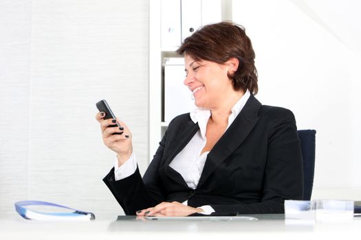 Businesswoman using her mobile at work sitting at her desk smiling in amusement at the text message she is reading on the screen A business woman checking her mobile phone and smiling after reading something.