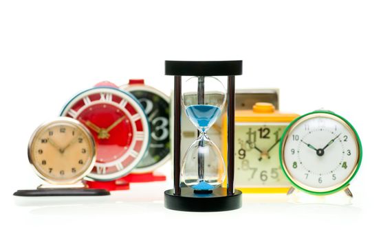 Sand timer with vintage mechanical wind-up alarm clocks on white background, shallow focus