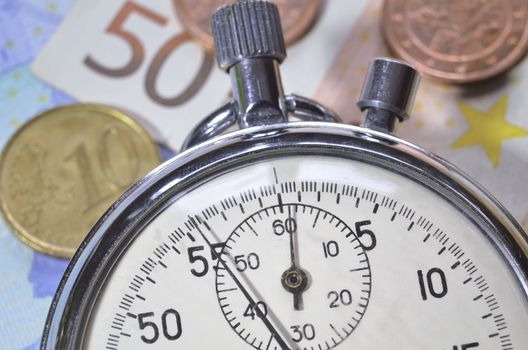 stopwatch lays over Euro coins and  banknotes; focus on watch arrow