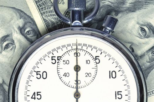 stopwatch lays over US dollar banknotes; focus on watch arrow